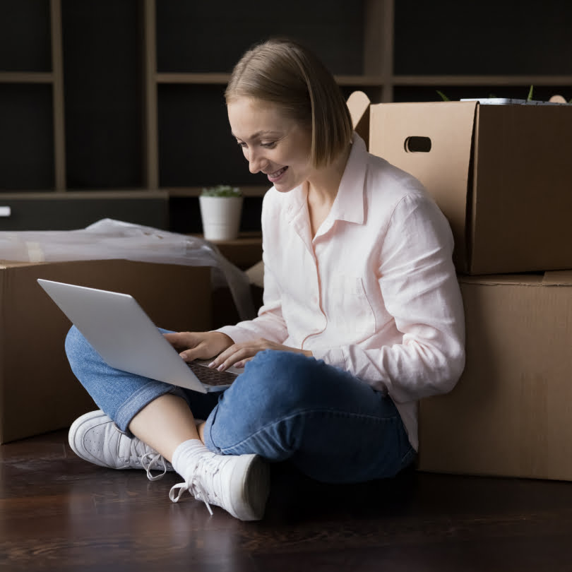 Nasmejana devojka sedi na podu sa laptopom naslonjena na kartonske kutije za selidbe.